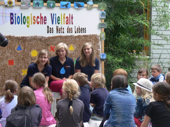 Das Bild zeigt eine Gruppe von Kindern, die aufmerksam einem Dreierteam von Erwachsenen zuhören