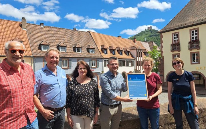 Das Bild zeigt sechs Personen, die vor dem Annweilerer Markplatz zusammenstehen. Im Hintergrund ist die Burg Trifels zu erkennen. Eine Person überreicht einer anderen einen Bilderrahmen, das ein Zertifikat enthält.