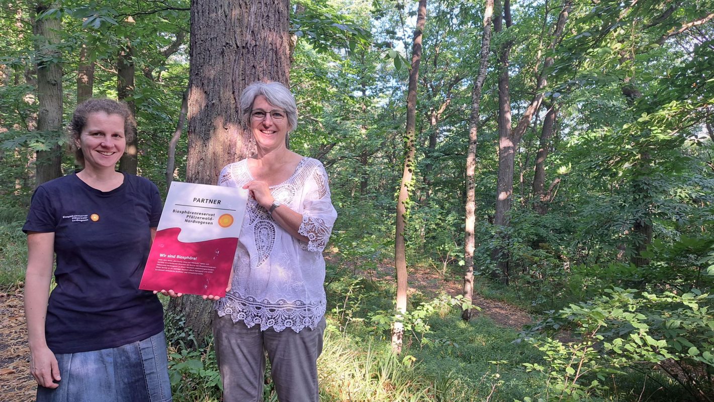 Dieses Bild zeigt die Direktorin des Biosphärenreservats, Friedericke Weber, und die Waldbademeisterin Britta Kollmann . Sie stehen im Wald und halten gemeinsam das Partner-Schild des Biosphärenreservats.
