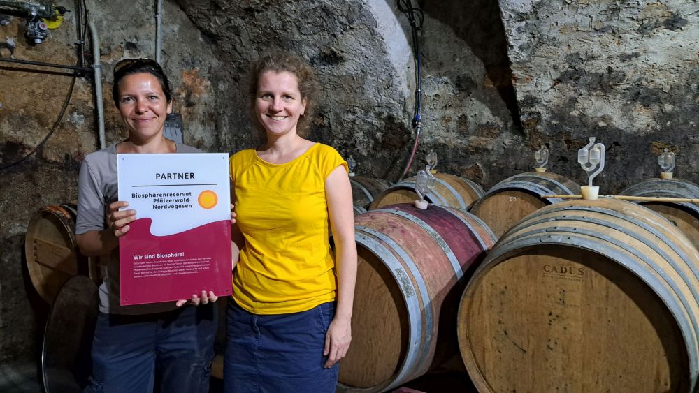 Es stehen zwei Personen in einem Weinkeller. Links steht Marie Adler mit der Auszeichnung zum Partnerbetrieb des Biosphärenreservats Pfälzerwald in der Hand. Rechts steht die Direktorin des Biosphärenreservats, Dr. Friedericke Weber.