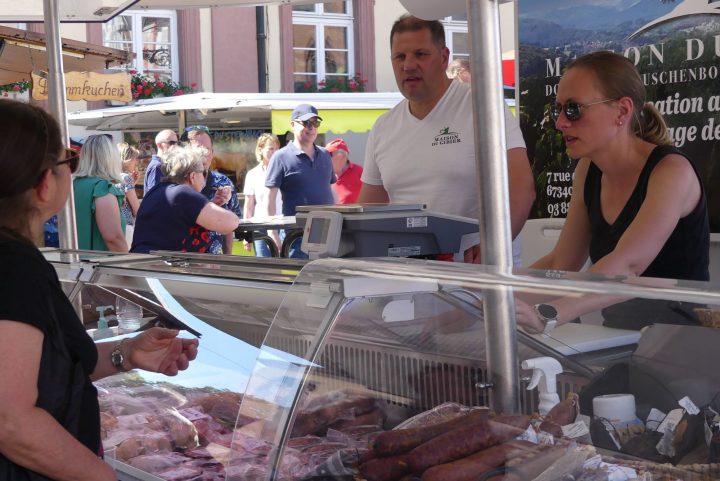 Eine Frau links im Bild steht vor einer Glastheke, in der Fleisch und Wurst ausgelegt ist. Hinter der Theke stehen zwei weitere Personen, die die Kundin bedienen.