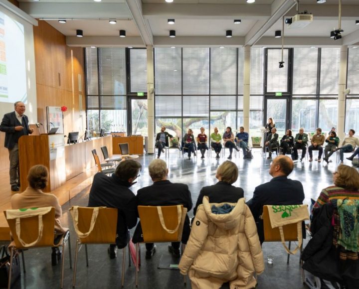 Das Bild zeigt einen Saal mit Personen, diie in einem Stuhlkreis sitzen. Links ein Podium mit einem Mann, der einen Vortrag hält.