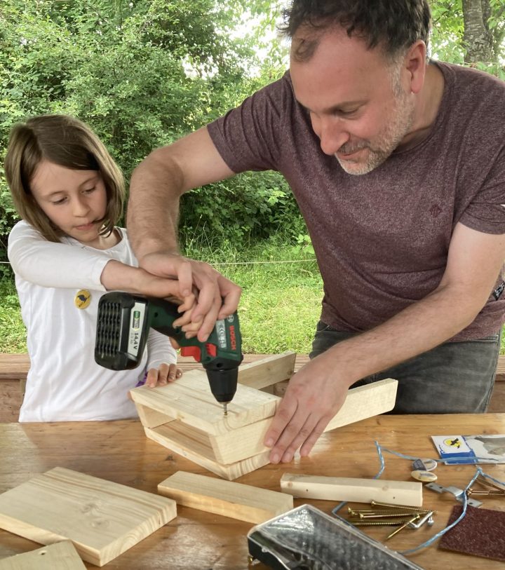 Das Bild zeigt ein etwa 8-jähriges Mädchen mit ihrem Papa. Die beiden bauen gemeinsam an einem Fledermauskasten.