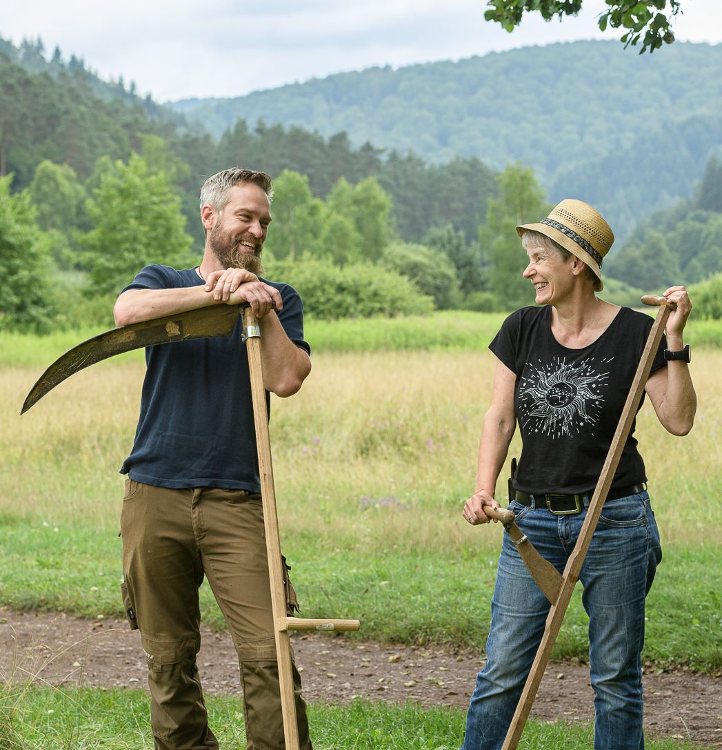Dieses Bild zeigt einen Mann und eine Frau. Sie haben Sensen in der Hand und stehen in einer typischen Landschaft des Biosphärenreservats mit Wiesen und Wald im Hintergrund.